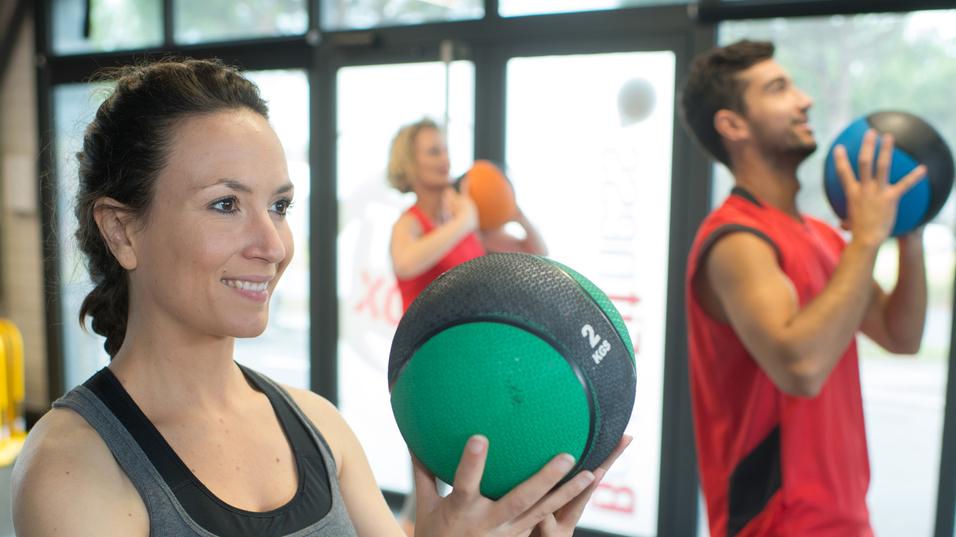 Drei Personen trainieren im Fitnessstudio mit Trainingsbällen; eine Frau hält einen grünen Medizinball.