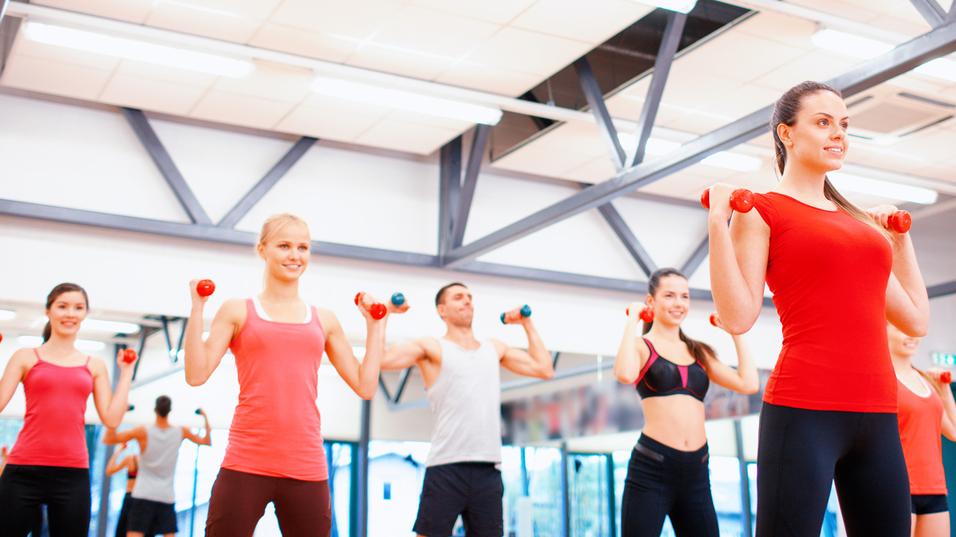 Gruppe von Menschen in Sportkleidung hebt Hanteln in einem Fitnessstudio mit Spiegeln und heller Beleuchtung.