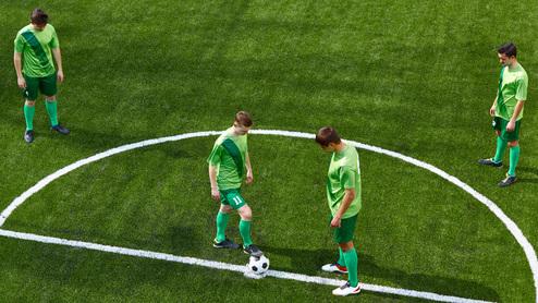 Vier Fußballspieler in grünen Trikots stehen auf einem Kunstrasenfeld, während ein Ball in der Mitte wartet.