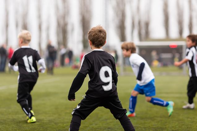 Kinder spielen Fußball auf einem Sportplatz; ein Spieler trägt ein schwarzes Trikot mit der Nummer 9.