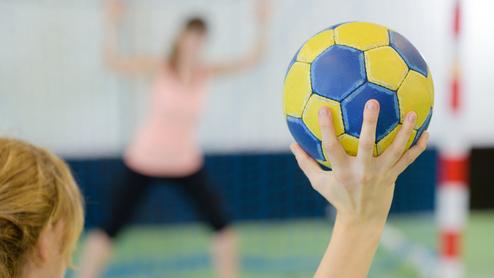 Nahaufnahme einer Hand, die einen gelb-blauen Handball hält, während eine Spielerin im Hintergrund positioniert ist.