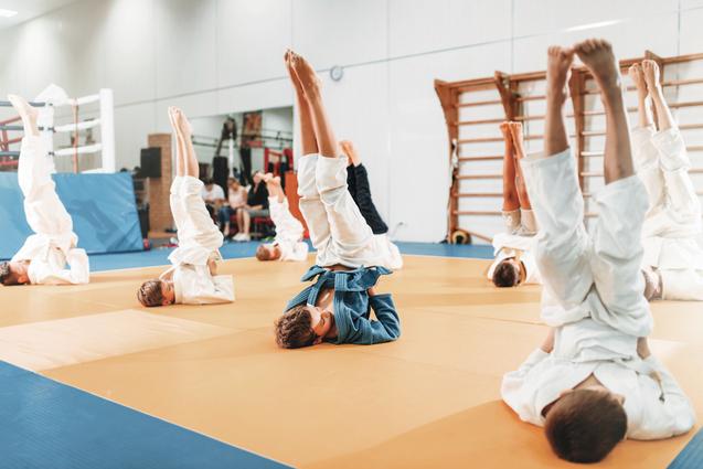 Gruppe von Kindern in Judoanzügen, die auf einer Matte Kopfstand-Übungen machen. Im Hintergrund sind Zuschauer sichtbar.