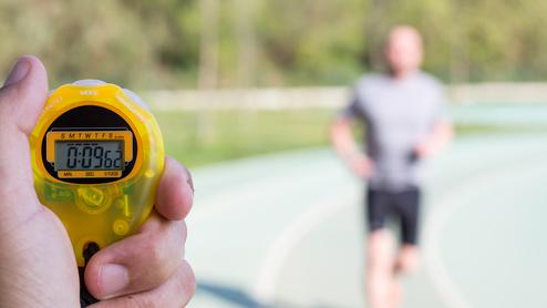 Hand hält eine gelbe Stoppuhr mit 00:08:66, während ein Jogger auf einer Laufbahn im Hintergrund läuft.