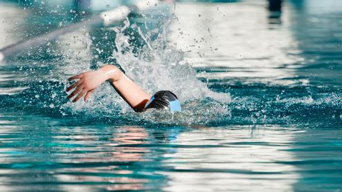 Schwimmerin in Schwarz mit blauer Badekappe zieht eine Bahn im Wasser, spritzt dabei Wasser auf.