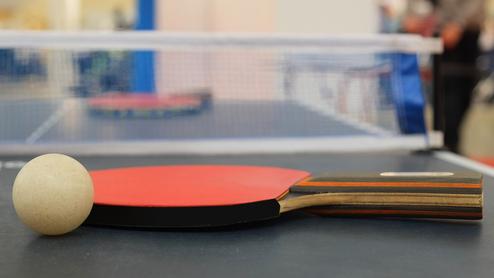 Nahaufnahme eines Tischtennisschlägers mit Ball auf einem Tischtennistisch mit Netz im Hintergrund.